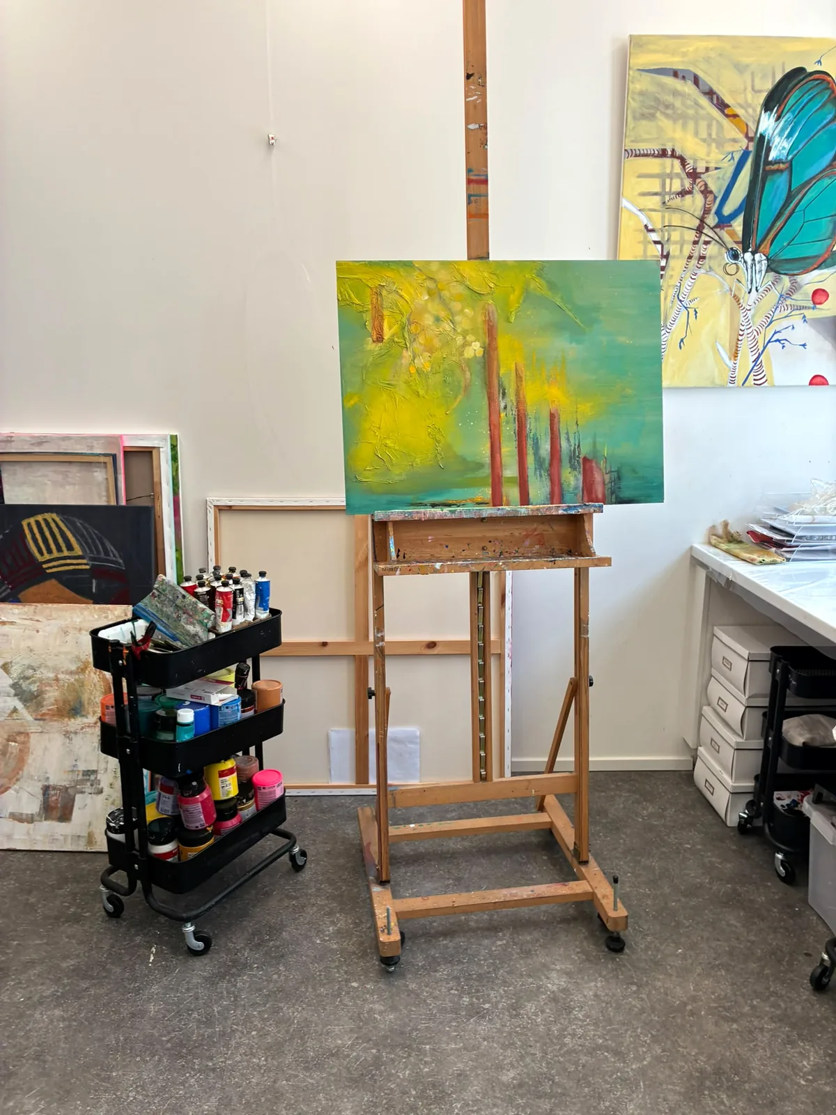 Het atelier: een houten ezel met een geelgroen doek, een karretje vol verf, een groot doek met een vlinder aan de muur.